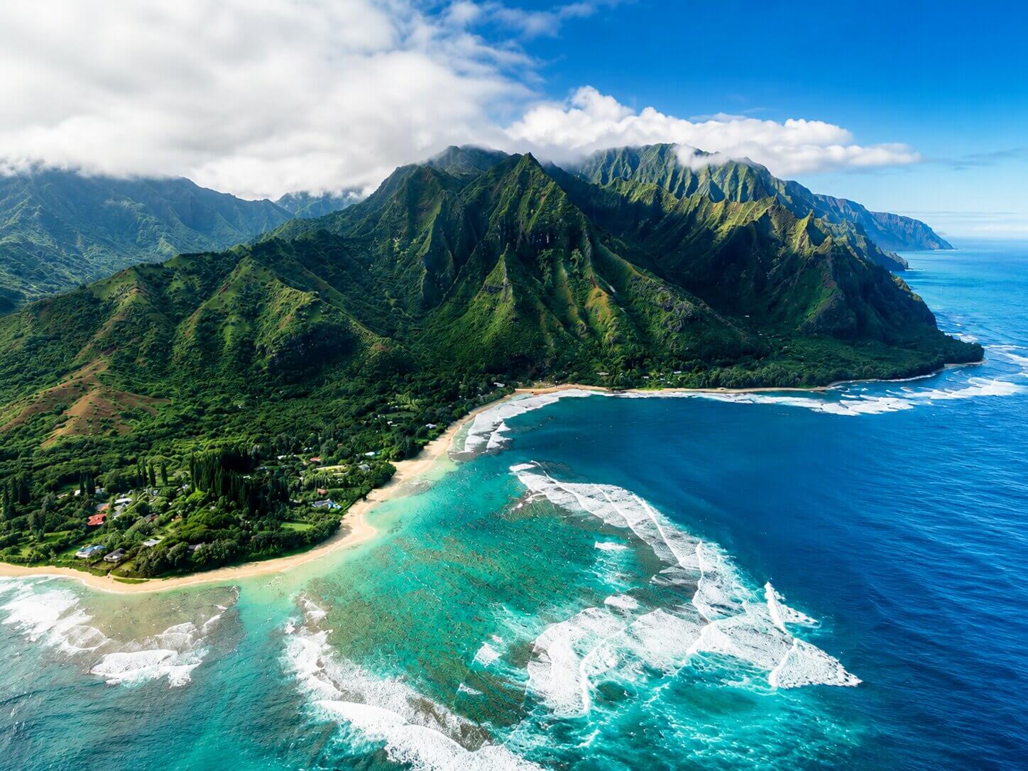 Aerial view of a Hawaiian island with green mountain ridges, sandy beach, and turquoise reef waters